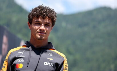 Close-up portrait of a young man in a grey and orange racing team hoodie with sponsor logos, outdoors in a mountainous area.