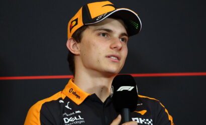 Young race driver in an orange cap and branded polo giving an interview, speaking into an F1 microphone.