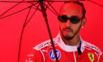 Man in a red Ferrari racing suit wearing dark sunglasses under a red umbrella, looking to the side.