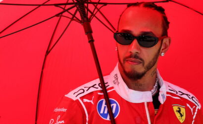 Man in a red Ferrari racing suit wearing dark sunglasses under a red umbrella, looking to the side.