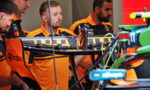 Pit crew in orange uniforms work on a race car in the garage, focusing on the rear wing and wiring connections.