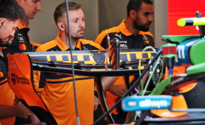 Pit crew in orange uniforms work on a race car in the garage, focusing on the rear wing and wiring connections.