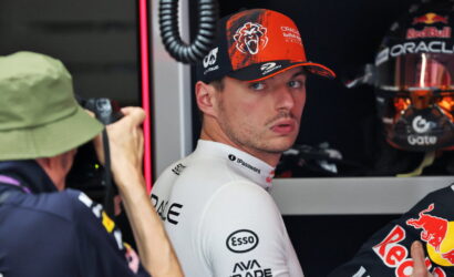 Race car driver in a white sponsor suit looks over his shoulder in a garage, photographer on the left taking a photo.