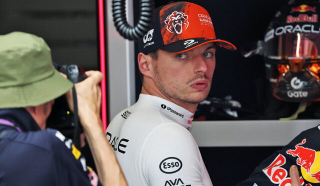 Race car driver in a white sponsor suit looks over his shoulder in a garage, photographer on the left taking a photo.