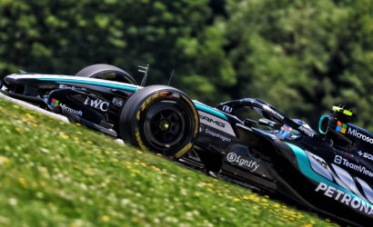 Black and teal Formula 1 car racing along a grassy slope with sponsor logos visible (Microsoft, IWC, Petronas) and a blurred green background.
