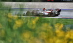 White and red Formula 1 car speeding on a wet track with sponsor banners in the background and blurred yellow flowers in the foreground.