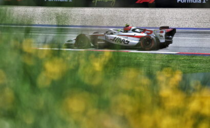 White and red Formula 1 car speeding on a wet track with sponsor banners in the background and blurred yellow flowers in the foreground.