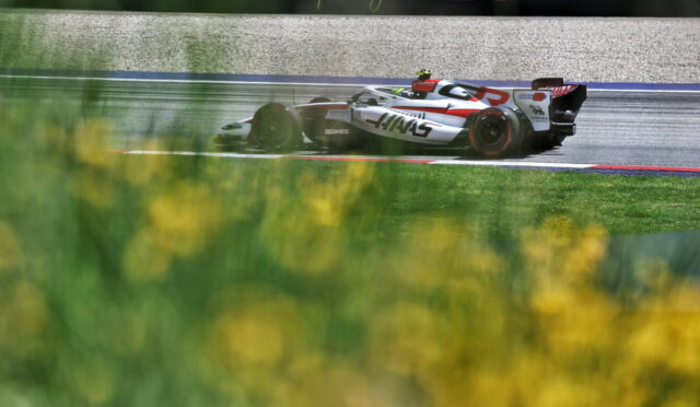 White and red Formula 1 car speeding on a wet track with sponsor banners in the background and blurred yellow flowers in the foreground.