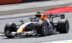Red Bull Racing Formula 1 car speeding through a corner on a race track, orange intake and sponsor logos visible on the bodywork.