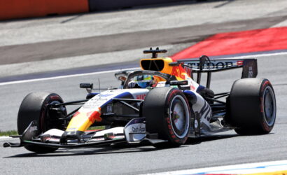 Red Bull Racing Formula 1 car speeding through a corner on a race track, orange intake and sponsor logos visible on the bodywork.