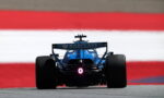 Blue Formula 1 car viewed from behind on a racetrack with a Barc lays sponsor rear wing visible and red-white-red barrier in the background