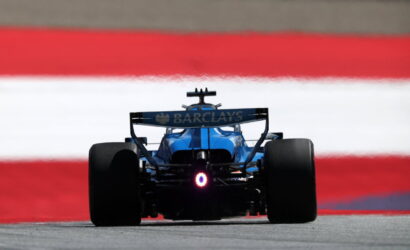 Blue Formula 1 car viewed from behind on a racetrack with a Barc lays sponsor rear wing visible and red-white-red barrier in the background