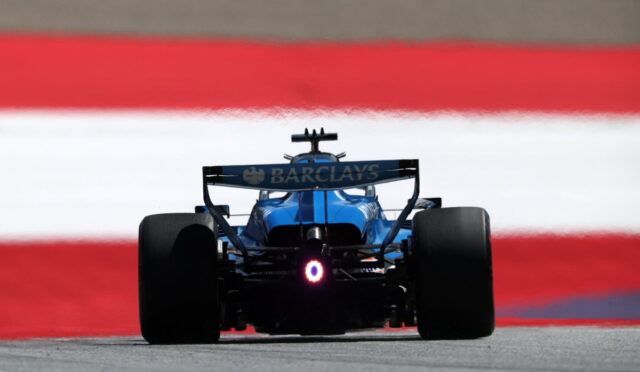 Blue Formula 1 car viewed from behind on a racetrack with a Barc lays sponsor rear wing visible and red-white-red barrier in the background