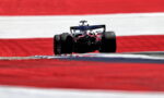 Rear view of a Formula 1 car racing on a track with red and white painted barriers in the background, speed blurred by the motion.