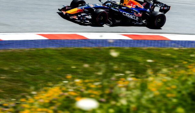 Red Bull Formula 1 car speeds along a race track; foreground shows blurred green grass and yellow flowers.