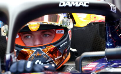 Close-up of a race car driver in a colorful helmet seated in a Formula 1 cockpit, DAMAC branding visible on the halo bar.