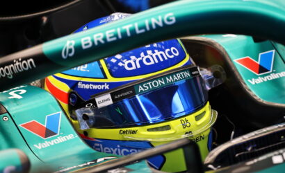 Close-up of a Formula 1 driver's helmet inside a teal Aston Martin car, featuring Aramco, Valvoline, and Breitling logos act as sponsors.