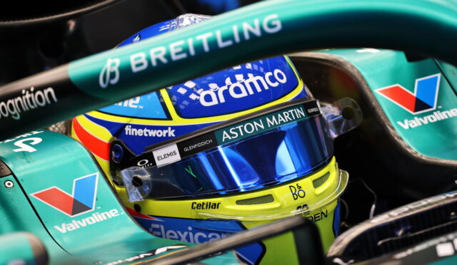 Close-up of a Formula 1 driver's helmet inside a teal Aston Martin car, featuring Aramco, Valvoline, and Breitling logos act as sponsors.