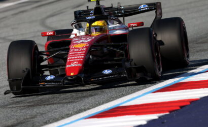Red Formula 1 race car drifts through a corner on a race track, sponsor logos visible on the nose and wings, driver in yellow helmet inside cockpit