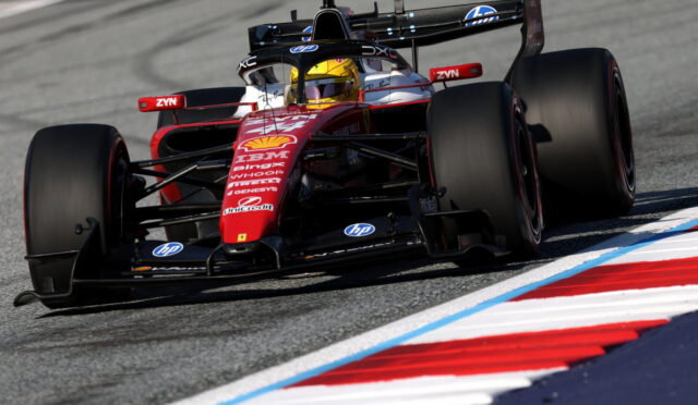 Red Formula 1 race car drifts through a corner on a race track, sponsor logos visible on the nose and wings, driver in yellow helmet inside cockpit