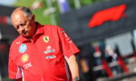 Man in a red Ferrari team polo with sponsor logos, looking down at his wristwatch at an outdoor racing event.