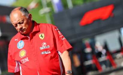 Man in a red Ferrari team polo with sponsor logos, looking down at his wristwatch at an outdoor racing event.