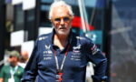 Older man with white hair and orange sunglasses, wearing a navy polo with multiple sponsor logos, standing outdoors at a racing event with a lanyard badge labeled PIT 2025.