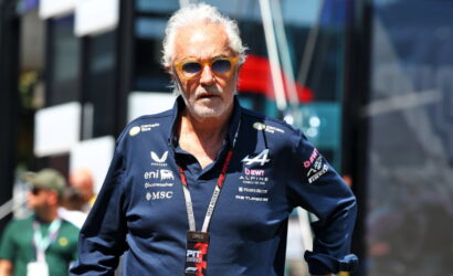 Older man with white hair and orange sunglasses, wearing a navy polo with multiple sponsor logos, standing outdoors at a racing event with a lanyard badge labeled PIT 2025.