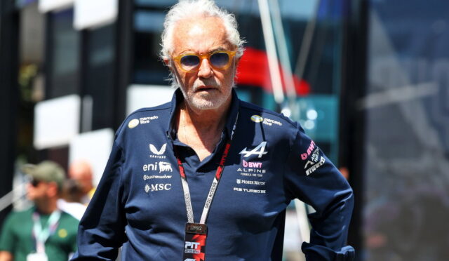 Older man with white hair and orange sunglasses, wearing a navy polo with multiple sponsor logos, standing outdoors at a racing event with a lanyard badge labeled PIT 2025.
