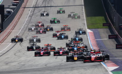 A crowded start of a Formula 1 race on a straight track, red and blue race cars lined up behind the leaders, tires smoking slightly as they accelerate. Left side shows pit lane and barriers, right side safety barriers and Lenovo banners.