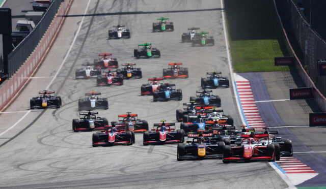 A crowded start of a Formula 1 race on a straight track, red and blue race cars lined up behind the leaders, tires smoking slightly as they accelerate. Left side shows pit lane and barriers, right side safety barriers and Lenovo banners.
