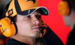 Man wearing a yellow racing headset and cap, focused in a pit area with a red background behind him.