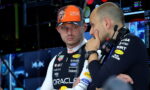 Two Formula 1 team members in a high-tech pit area; one wears an orange Oracle Red Bull Racing cap and white racing suit with sponsor logos, listening to the other who wears a headset.