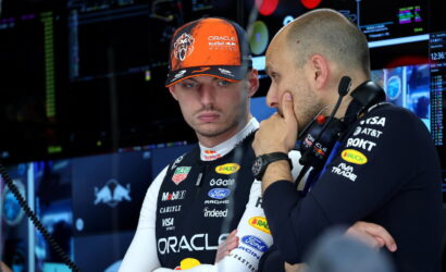 Two Formula 1 team members in a high-tech pit area; one wears an orange Oracle Red Bull Racing cap and white racing suit with sponsor logos, listening to the other who wears a headset.