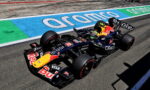 Red Bull Racing Formula 1 car on a race track with sponsor decals visible on the body and tires kicked up slight dust nearby the wall art behind it.