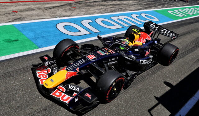 Red Bull Racing Formula 1 car on a race track with sponsor decals visible on the body and tires kicked up slight dust nearby the wall art behind it.
