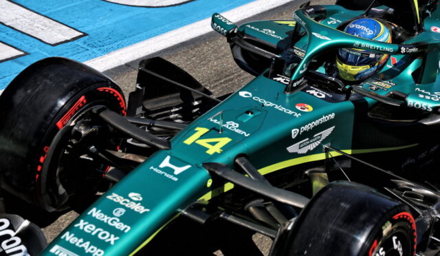 Teal Aston Martin F1 car #14 on the track, close-up of the front section with sponsor logos and a visible driver helmet.