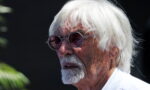 Older man with white hair and round sunglasses, looking to the left against a dark background.