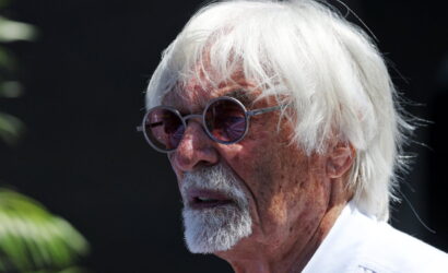 Older man with white hair and round sunglasses, looking to the left against a dark background.