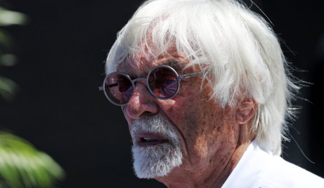 Older man with white hair and round sunglasses, looking to the left against a dark background.