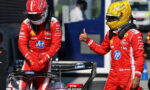 Two Ferrari Formula 1 drivers in red racing suits stand beside a race car in the pit lane; the driver on the right gives a thumbs-up.