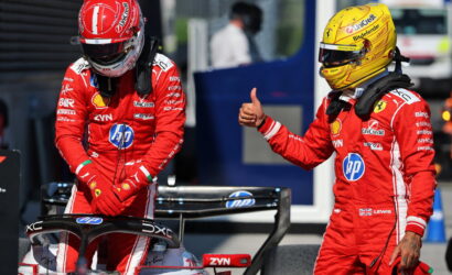 Two Ferrari Formula 1 drivers in red racing suits stand beside a race car in the pit lane; the driver on the right gives a thumbs-up.