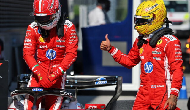 Two Ferrari Formula 1 drivers in red racing suits stand beside a race car in the pit lane; the driver on the right gives a thumbs-up.