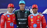 Three Formula 1 drivers pose on a podium; center driver in black Petronas suit and blue cap, flanked by Ferrari drivers in red suits with hands on shoulders.