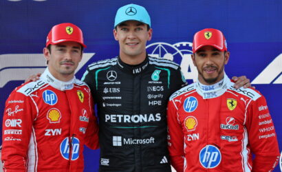 Three Formula 1 drivers pose on a podium; center driver in black Petronas suit and blue cap, flanked by Ferrari drivers in red suits with hands on shoulders.