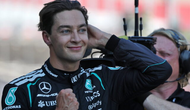 Male race car driver in a Mercedes Petronas suit smiles and touches his ear while a photographer films in the background.