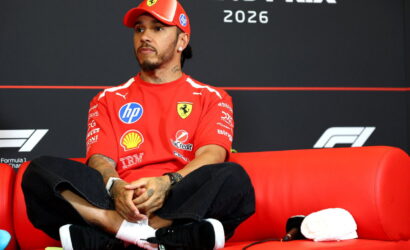 Ferrari Formula 1 driver seated on a red couch at a press event, wearing a red cap and shirt with sponsor logos and a 2026 backdrop.