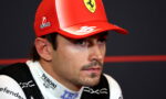 Close-up of a race car driver in a red Ferrari cap during a press conference, with sponsor logos visible on cap and suit.