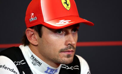 Close-up of a race car driver in a red Ferrari cap during a press conference, with sponsor logos visible on cap and suit.