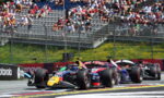 Three Formula 1 race cars cornering on a track as a crowded grandstand watches.